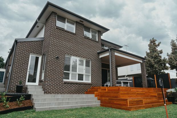 Exterior view of a two-storey suburban brick house with a low-pitched roof, white louvred window shutters, a covered entry or carport-like structure, grey tiled steps, and a prominent raised timber deck and stair platform at the front. The property sits within a landscaped residential setting under an overcast sky.