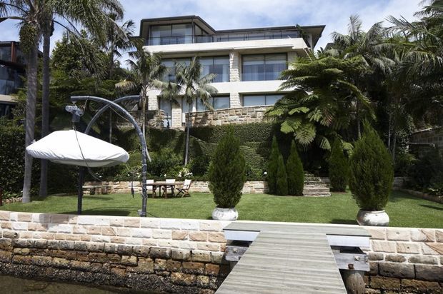 Exterior view of a multi-storey contemporary waterfront residence with broad horizontal glazing, balconies, pale stone cladding and landscaped grounds. A timber jetty leads toward the property, with palms, clipped hedges, conifers and a covered outdoor swing or daybed in the foreground.