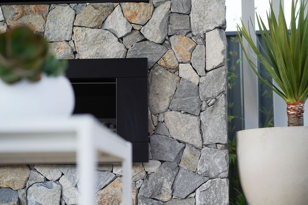 Close-up interior view of a substantial irregular natural stone feature wall surrounding a dark built-in fireplace or media opening. A white table or console is softly blurred in the foreground, while a tall potted architectural plant stands beside grey-framed glazing at right. The image has a refined, contemporary natural-material aesthetic.