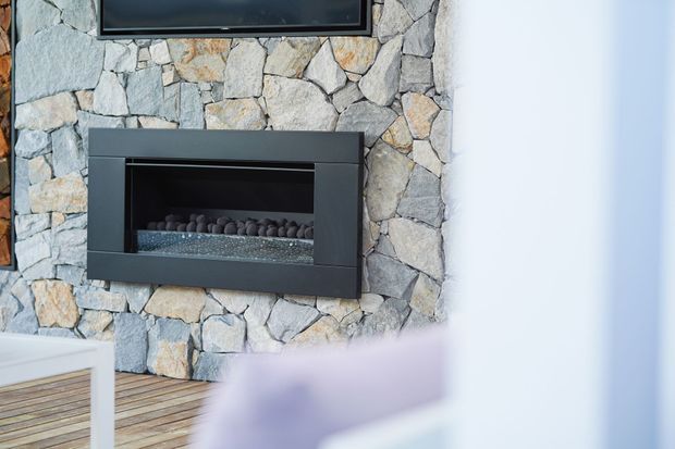 Close-up interior view of a contemporary electric fireplace set into a rugged irregular natural-stone feature wall, with a television mounted above. Pale timber flooring and a partial white built-in element are visible at the lower left.