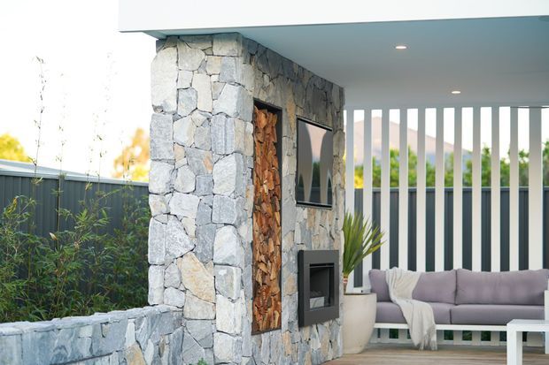 Covered outdoor entertaining area with a contemporary flat roof, pale vertical privacy screens, and a substantial irregular-stone-clad fireplace wall. The wall incorporates a stacked firewood niche, television, and built-in fireplace, with a modern upholstered sofa and planters arranged on a timber-toned deck. A dark boundary fence and landscaped planting are visible beyond.