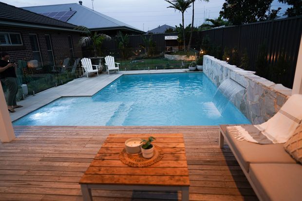 A residential outdoor entertaining area with a rectangular swimming pool, timber decking, outdoor lounge seating, and a raised stone-clad pool wall. The pool is bordered by glass fencing and planted landscaping, with neighbouring homes visible beyond the dark privacy fence. The scene has warm evening lighting and a relaxed resort-like atmosphere.
