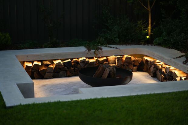 A contemporary sunken outdoor fire pit set into a pale concrete terrace, with a circular black metal fire bowl surrounded by neatly stacked firewood and warm concealed lighting. Dense planting and a dark vertical fence form the background, with lawn in the foreground.