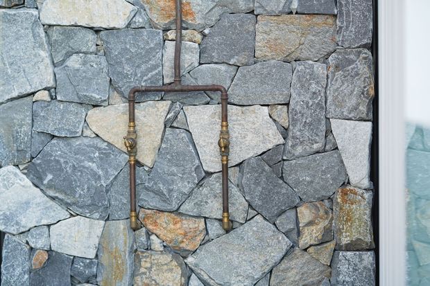 Close-up of a rustic interior wall clad in irregular natural grey stone, with exposed copper-toned plumbing and brass fittings mounted across the surface. A narrow glimpse of a pale tiled or stone adjacent area appears at the right edge.