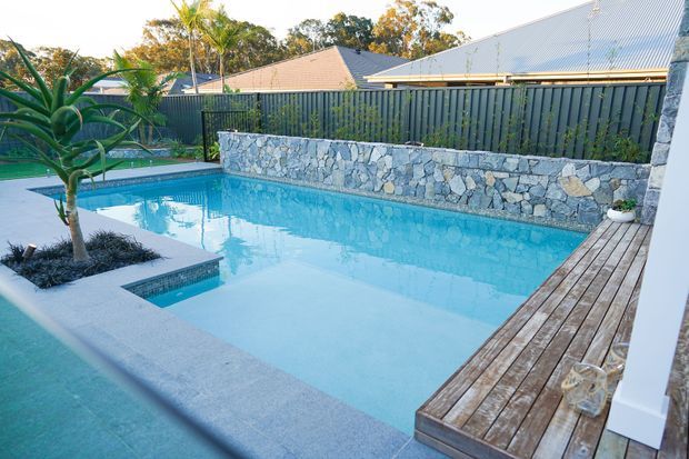 A bright residential outdoor pool area with a turquoise swimming pool, integrated shallow ledge and tiled step, surrounded by pale concrete coping and a timber deck. A low irregular natural-stone retaining wall and dark vertical privacy fencing border the pool, with tropical planting and suburban rooftops beyond.