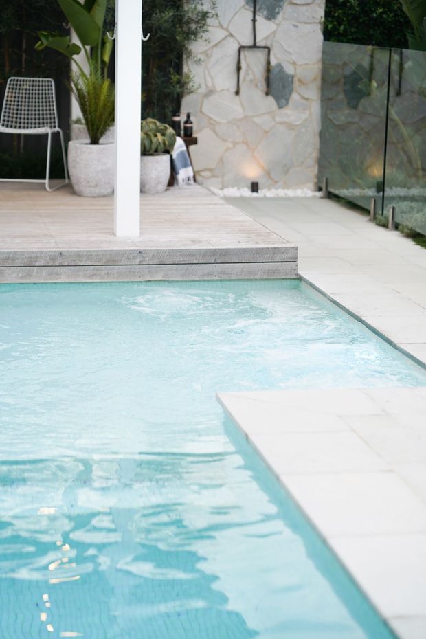 A bright contemporary outdoor pool area with pale aqua water, white paving, and a light timber pool deck. A glass privacy screen runs along one side, while a natural irregular stone feature wall, tropical plants, potted greenery, and a white outdoor chair create a relaxed resort-like setting.