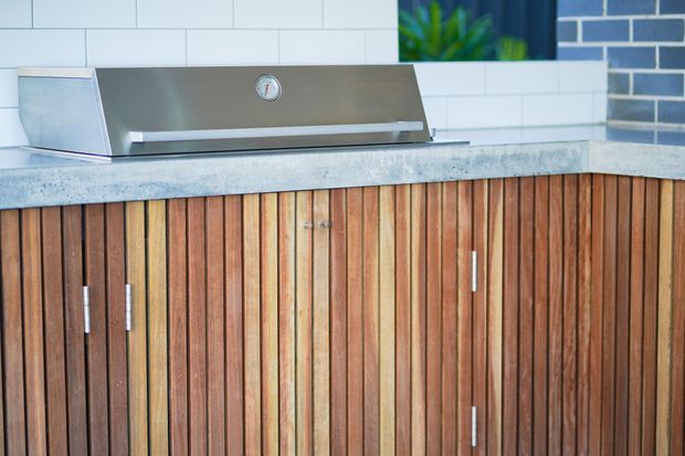 Close-up of a built-in outdoor kitchen barbecue set into a timber-clad cabinet. The cabinet has vertical narrow timber battens and slim metal handles, topped by a mottled grey concrete benchtop. Behind it is a white rectangular tiled splashback with a dark blue-grey brick accent wall and greenery visible above.