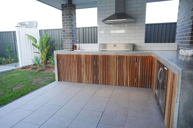 A covered outdoor kitchen and barbecue area with a large tiled floor, stainless-steel grill and rangehood, grey tiled and rendered walls, dark grey metal fencing, and distinctive vertical timber-slatted cabinetry. The space opens onto a small landscaped yard with tropical planting.