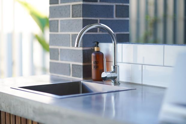 Close-up of a contemporary kitchen sink with a tall chrome gooseneck tap set into a pale stone benchtop. The backsplash combines dark grey brick-pattern tiles with white rectangular subway tiles, and a soap dispenser sits beside the tap. Bright natural light enters from the left.