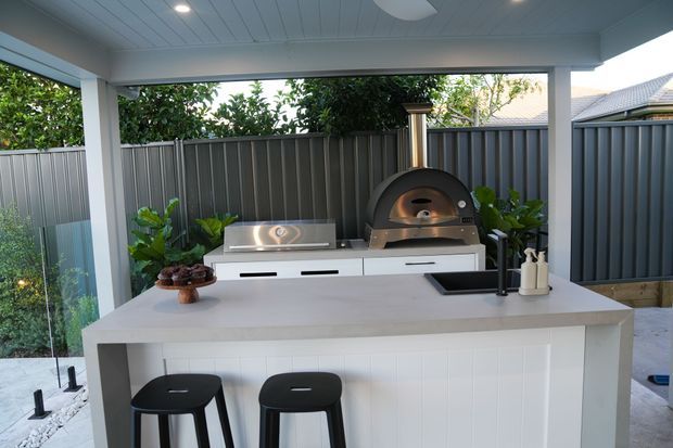 A covered outdoor kitchen and entertaining area with a large white island bench, two black stools, built-in barbecue and wood-fired pizza oven. The space is enclosed by dark corrugated metal fencing and landscaped with greenery, with a white-lined ceiling and partial glass screening.