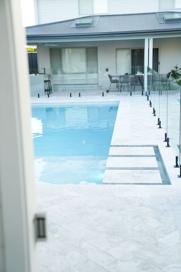 A bright modern courtyard pool viewed from through a doorway, with a pale tiled terrace surrounding a rectangular swimming pool. Frameless glass balustrades with dark posts line the pool edge, and stepping slabs cross a shallow section. A contemporary white building with a grey metal roof, skylights, large glazing and an outdoor dining setting forms the background.