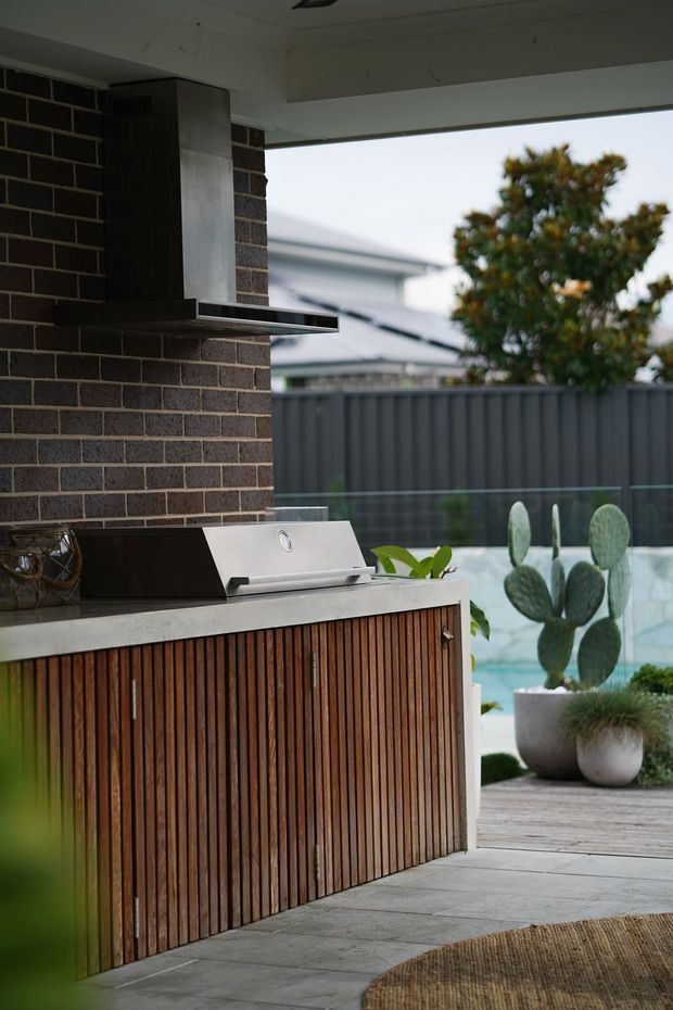 A covered outdoor kitchen beside a pool, featuring a built-in stainless-steel barbecue and rangehood, dark brickwork, a pale stone benchtop, and vertical timber-slatted cabinetry. Glass pool fencing, potted cactus plants, and a timber deck are visible beyond.