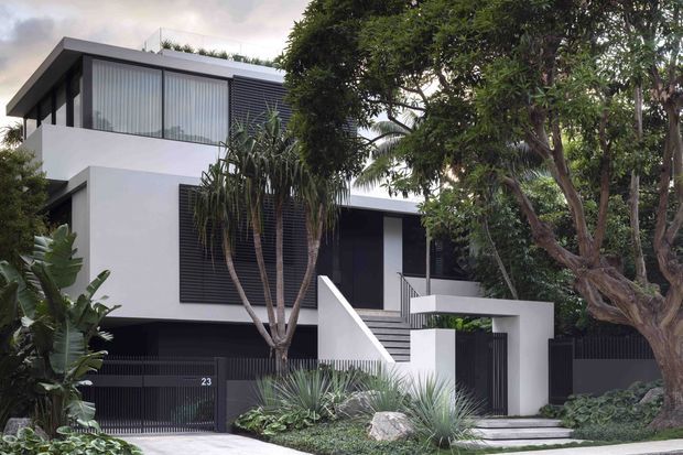 Street-facing view of a contemporary multi-storey residence with a crisp white rendered facade, dark horizontal screens and fencing, extensive glazing, flat rooflines, and a recessed entry reached by steps. Tropical and sculptural landscaping frames the building.