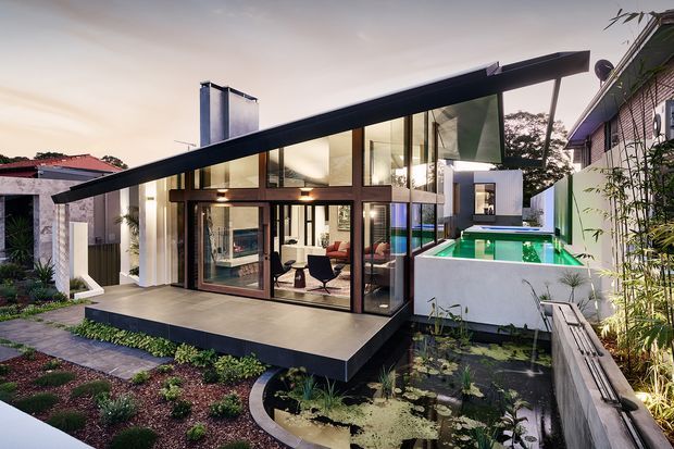Exterior view of a contemporary glass-walled house with a dramatic single-slope roof, dark structural framing, elevated terrace, and illuminated turquoise swimming pool. A landscaped garden with a small pond surrounds the home, while the interior living and dining areas are visible through the glazing.