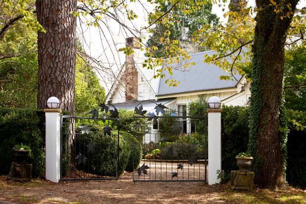 A leafy residential property viewed from the street through a decorative metal entrance gate. The home has a steep gabled roof, light exterior walls, a prominent brick chimney, and a landscaped garden enclosed by hedges and mature trees. White gate pillars are topped with spherical lights, and bird silhouettes decorate the gate.