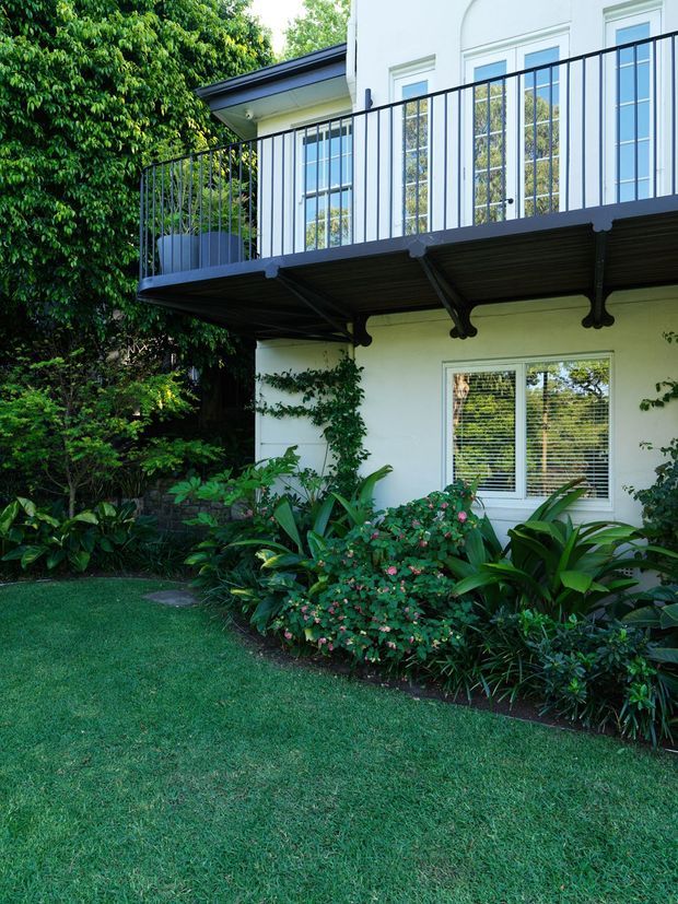 Exterior view of a light-painted multi-level house with a projecting upper balcony, dark metal balustrade, and exposed dark structural supports beneath the balcony. White-framed windows reflect surrounding greenery. A lush landscaped garden with dense planting and manicured lawn occupies the foreground.