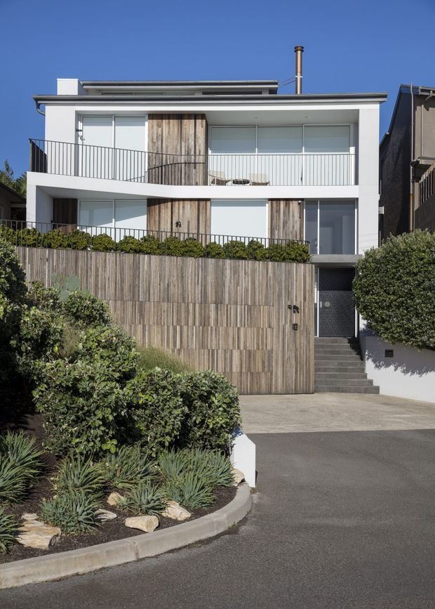 Street-facing exterior view of a contemporary multi-storey residence with a flat roof, white rendered facade, broad glazed openings, slim metal balustrades, and vertical timber cladding used across the front fence and selected wall sections. A recessed entry is reached by steps beside landscaped planting and a driveway.