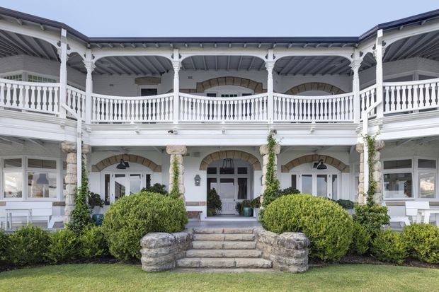 Symmetrical two-storey courtyard-facing building facade with white painted columns, ornate balustrades, covered verandas, arched stone-trimmed openings, grey metal roofing, and landscaped hedges around a central staircase. The architecture has a refined traditional character with strong formal detailing.