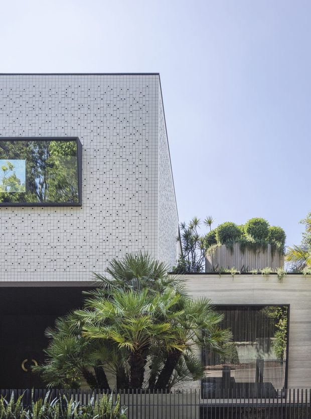 A contemporary multi-level building facade photographed from outdoors. The upper volume is clad in small light-grey patterned tiles or panels with a large dark-framed window reflecting trees. A lower section has dark textured cladding, broad timber-toned horizontal paneling, and large glazed openings. Tropical and sculptural planting, including palms and yucca-like plants, occupies the foreground and roof terrace. The composition emphasizes clean geometric forms, strong horizontal and vertical lines, and a restrained grey, black, and green palette under a clear sky.