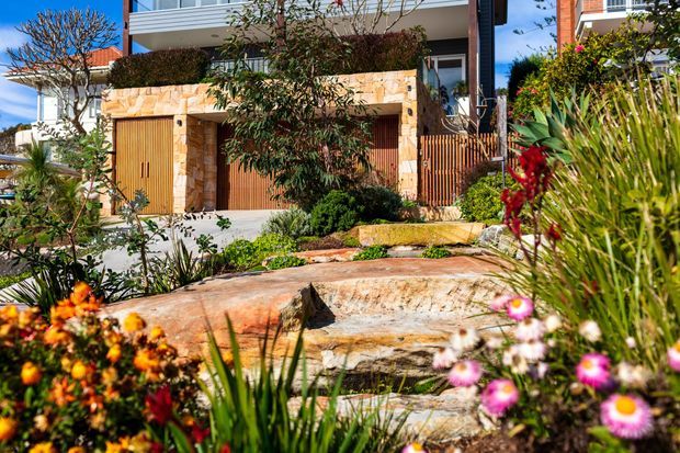 A landscaped residential frontage with a contemporary multi-storey house behind layered sandstone retaining walls. The facade combines warm stone cladding, timber garage or entry doors, vertical timber screening, white balcony structures, and extensive native-style planting with flowering groundcover.