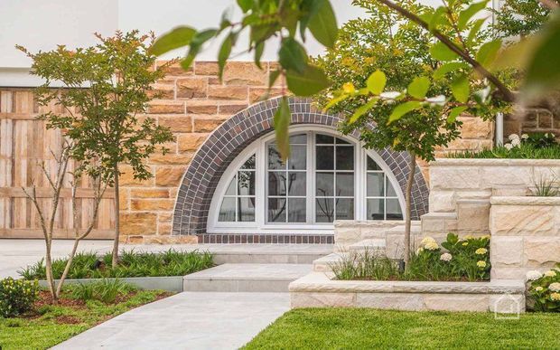 Landscaped front approach to a stone-clad building, featuring a distinctive arched window with dark masonry voussoirs, pale stone retaining walls and steps, timber double gates, and carefully planted garden beds.