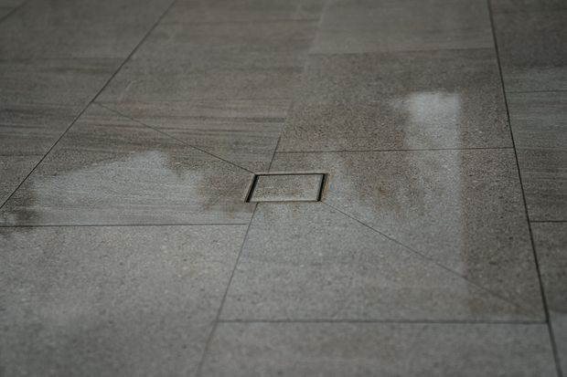 Close-up of a grey tiled floor with large-format rectangular tiles, visible grout lines, and a centrally positioned square floor drain or access cover. The surface has a subtle mottled stone-like appearance and a slightly reflective finish.