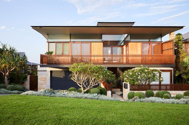 Two-storey contemporary residential home with a broad low-pitched roof, projecting upper-level balcony, extensive glazing, warm timber screens and balustrades, and a stone-accented ground floor. The landscaped frontage includes a manicured lawn, layered planting and sculpted shrubs.