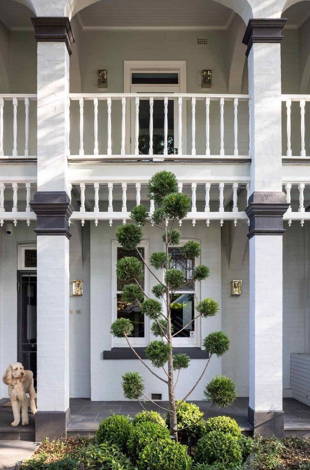 Front exterior view of a grand two-storey residence with a symmetrical colonnaded facade, repeated arched openings, white balustrades, dark trim accents, and pale rendered walls. A manicured courtyard garden with clipped shrubs and a sculpted tree occupies the foreground, with a small dog near the entrance.