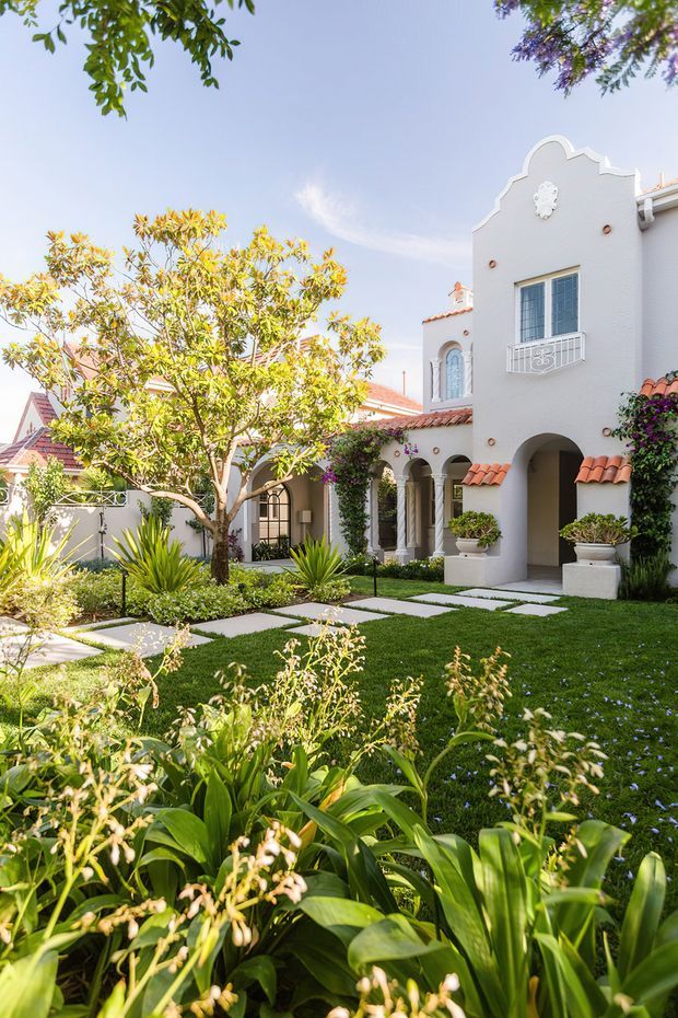 Bright exterior view of a refined white stucco residence with a decorative stepped gable, arched entry openings, tiled awnings, wrought-style balcony railing, and terracotta roof details. A formal landscaped garden with clipped lawn, paved stepping paths, raised planters, flowering vines, and lush planting fills the foreground.