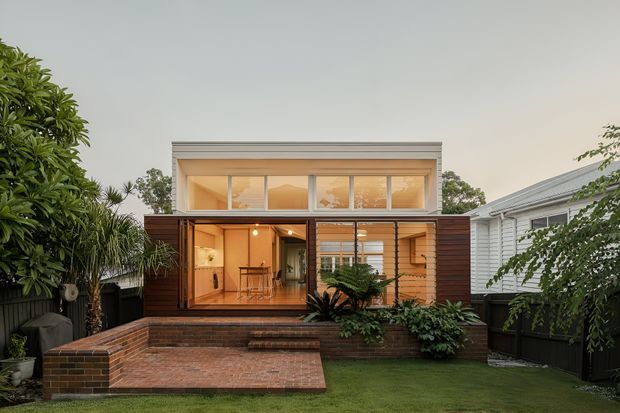 Rear exterior view of a compact two-storey residential extension with warm timber cladding, broad glazed openings, and a raised brick-paved terrace with steps leading to a small lawn. The upper level has a simple projecting box form with horizontal windows, while lush planting and neighbouring residential structures frame the scene.