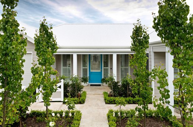 Front exterior of a single-storey light-colored home with a symmetrical entry facade, white weatherboard cladding, a pitched corrugated metal roof, shuttered windows, and a bright turquoise-blue front door. A straight paved path leads through a formal garden with clipped young trees and low hedging.