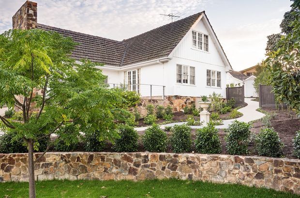 Exterior view of a white rendered, gable-roofed house with a prominent stone chimney, multi-pane windows, dark tiled roofing, and a landscaped front garden. A low natural-stone retaining wall, clipped hedges, paved walkway, and timber fencing define the approach to the home.
