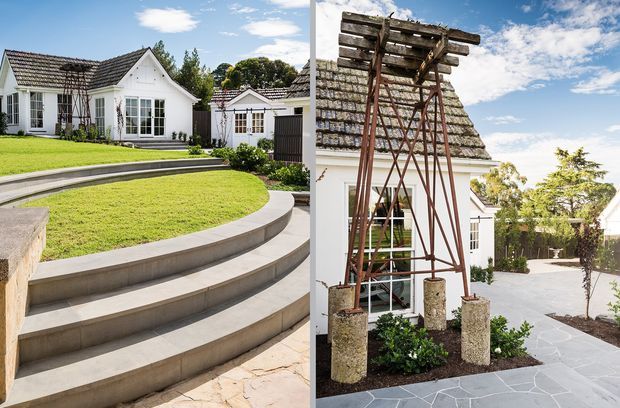 Exterior view of a white, gabled cottage-style residence with a tiled pitched roof, dark-framed windows, manicured lawn, curved concrete retaining steps, and a paved courtyard. A tall rustic timber pergola or trellis structure with climbing plants stands prominently in the foreground.