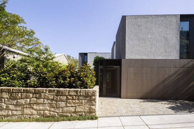 Street-facing exterior of a contemporary residence with a rectilinear, flat-roofed form. The facade combines dark textured rendered walls with vertical dark timber or wood-look cladding, a recessed entry, large glazing, and a pale stone block retaining wall. A paved driveway and footpath occupy the foreground, with established landscaping screening the neighboring structures.