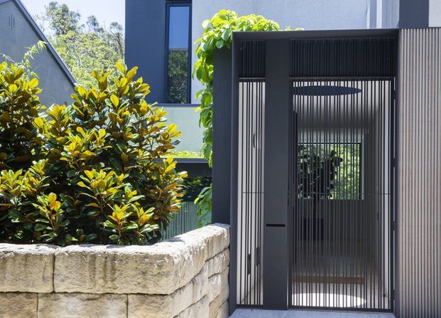 Contemporary residential entry gate in dark metal with vertical slatted detailing, set beside a rough-cut light stone retaining wall and dense landscaped planting. A pale rendered building facade with dark-framed glazing is visible behind the gate.