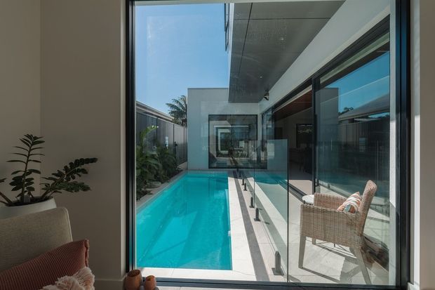 View from a contemporary interior through full-height glazing toward a narrow turquoise swimming pool in a private courtyard. The composition shows crisp white and grey architectural surfaces, dark-framed glazing, a covered outdoor walkway with slim balustrade, and a wicker lounge chair. A glimpse of indoor seating and a potted plant appears in the foreground.