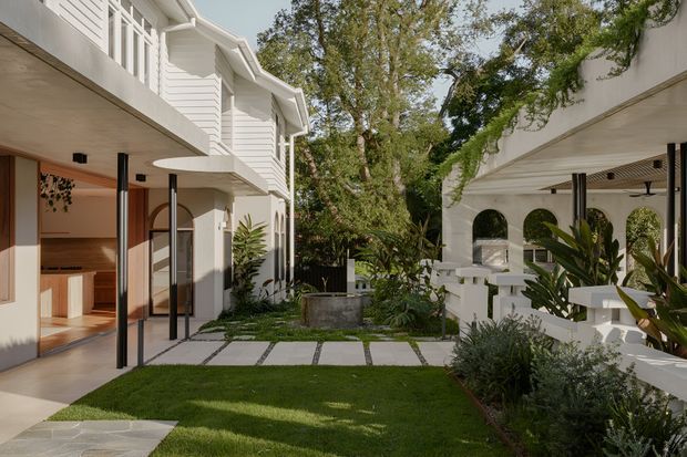 A refined courtyard between two white rendered and weatherboard-clad residential wings. The space features a clipped lawn, large square stepping stones, lush tropical planting, covered outdoor areas supported by slender dark posts, arched openings, and white balustrades. The left building has a contemporary coastal character with a visible kitchen or living interior opening onto the courtyard.