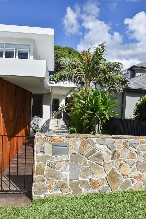 Bright exterior view of a contemporary two-storey residence with clean white rendered volumes, a projecting upper balcony, warm vertical timber cladding, and a timber entry walkway. A low irregular natural-stone boundary wall fronts the property, with lush tropical planting and a neighbouring modern home visible behind it.