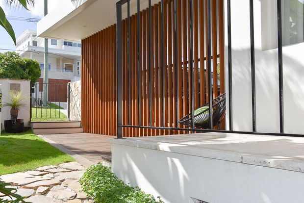 A bright contemporary residential entrance and terrace viewed from the landscaped front yard. The facade combines white rendered surfaces, extensive dark-framed glazing, and a prominent screen of closely spaced warm timber vertical battens. A timber walkway, raised concrete terrace, stone stepping path, lawn, and small planting beds lead toward the entry, with neighbouring residential buildings visible beyond.