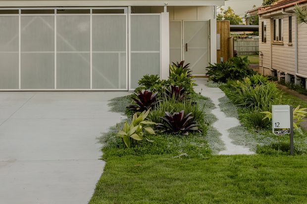 A landscaped residential frontage with a broad pale concrete driveway, translucent-panel garage doors, and a narrow gravel pedestrian path leading to a side entry. Layered tropical-style planting with grasses and burgundy foliage borders the path. A small raised timber-clad dwelling or neighbouring structure is visible at right, with a simple mailbox marked 12 in the foreground.