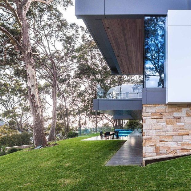 Contemporary multi-level house set among mature trees, featuring projecting dark-clad upper volumes, timber-lined soffits, expansive glazing, a glass-balustraded balcony, and a natural stacked-stone lower facade. A manicured lawn, paved terrace with outdoor dining, and swimming pool are visible.