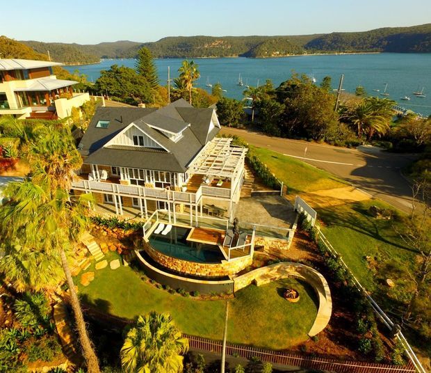 Aerial view of a multi-storey coastal residence with grey pitched roofing, white weatherboard-style exterior detailing, broad verandas and a partially covered outdoor terrace. The property features a curved swimming pool, landscaped lawn and tropical planting, with a road and sheltered bay visible beyond.