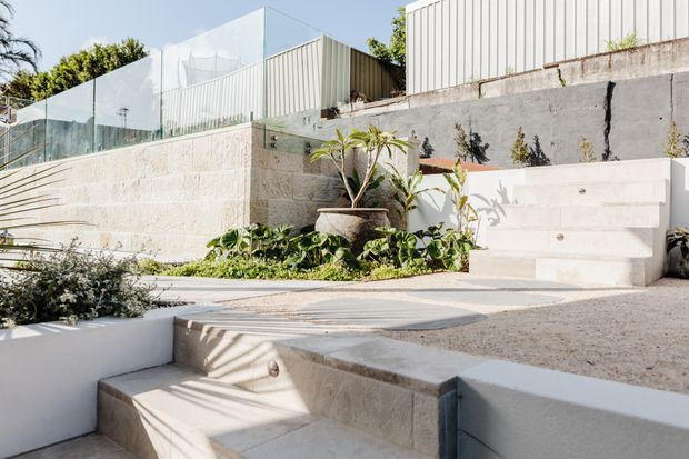 A bright, modern stepped outdoor terrace or courtyard with pale concrete and stone retaining walls, integrated stairs, landscaped planting beds, and a clear glass balustrade. White vertical-cladding structures sit above the terraced garden, creating a clean, sculptural residential exterior.
