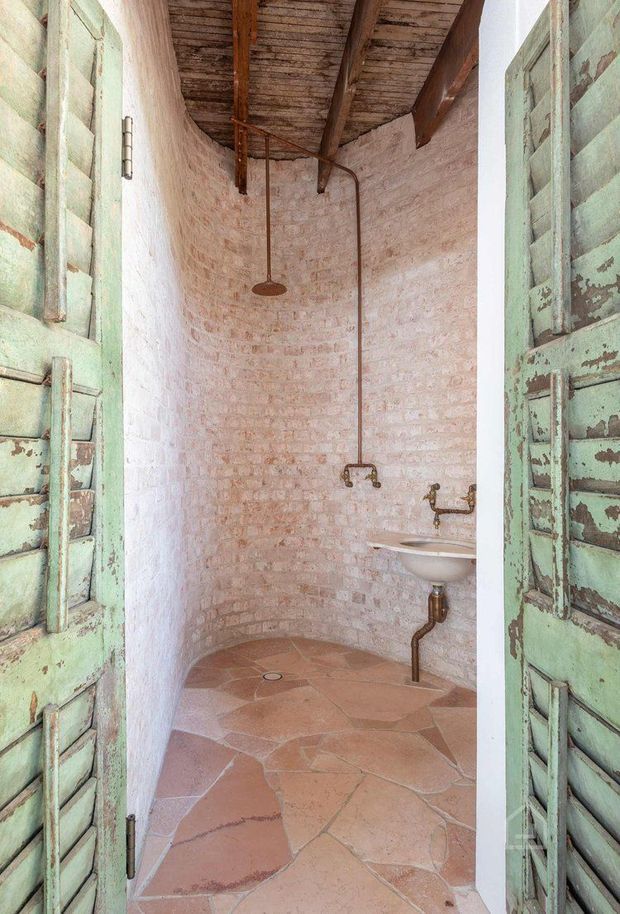 A narrow rustic bathroom viewed through distressed pale-green timber louvered doors. The space features exposed whitewashed brick walls, an aged timber-beam ceiling, exposed copper plumbing, a wall-mounted white ceramic basin, and irregular terracotta or natural stone floor tiles. A rainfall shower is mounted at the rear, creating an earthy, weathered, heritage-inspired atmosphere.