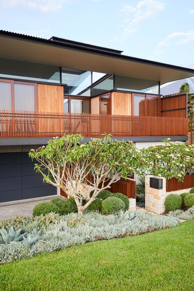 Contemporary two-storey residential facade with a broad low-pitched roof, extensive glazing, warm timber wall cladding and vertical timber privacy screening. A dark sectional garage door sits beneath the upper level, while a carefully designed front garden features clipped shrubs, sculptural planting, stonework and lawn.