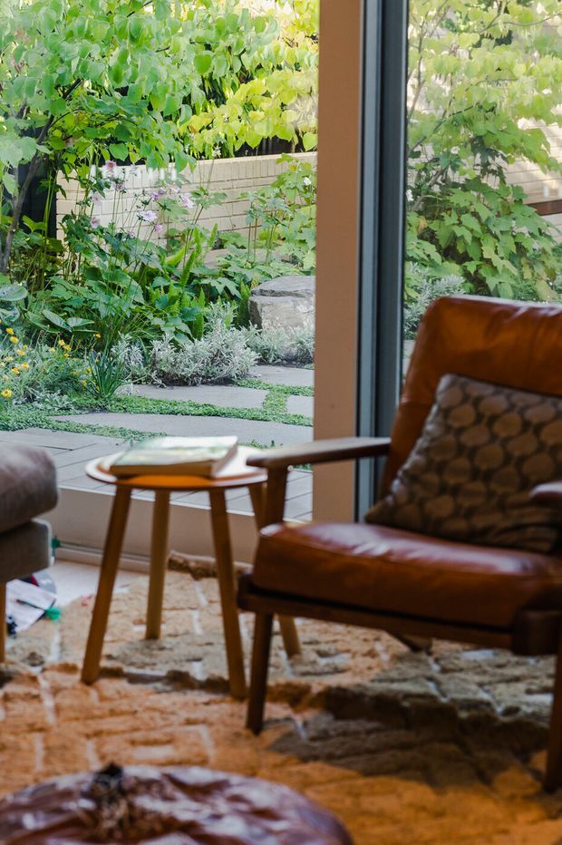A warm, mid-century-inspired sitting area beside expansive floor-to-ceiling glazing, furnished with a brown leather lounge chair, patterned cushion, timber side table, textured rug, and leather ottoman. The glazing opens views to a lush planted courtyard garden with a paved path and low masonry retaining wall.
