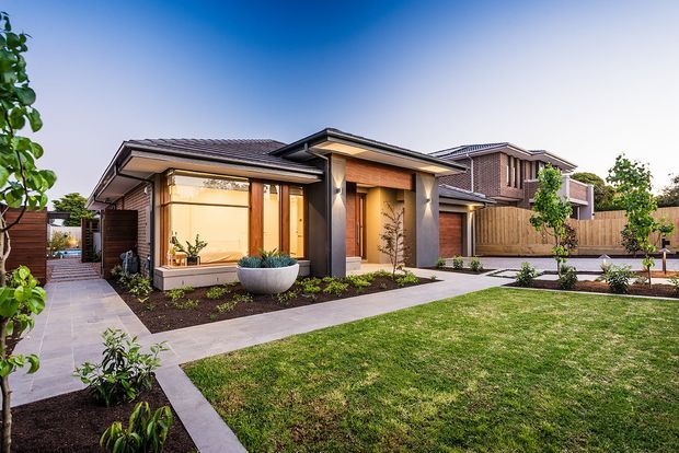 A contemporary single-storey suburban residence viewed from the front garden, with a low-pitched tiled roof, rendered and dark feature-clad walls, warm timber entry detailing, broad glazed openings, landscaped planting beds, paved paths, and a manicured lawn. A larger two-storey wing is visible behind the main frontage.