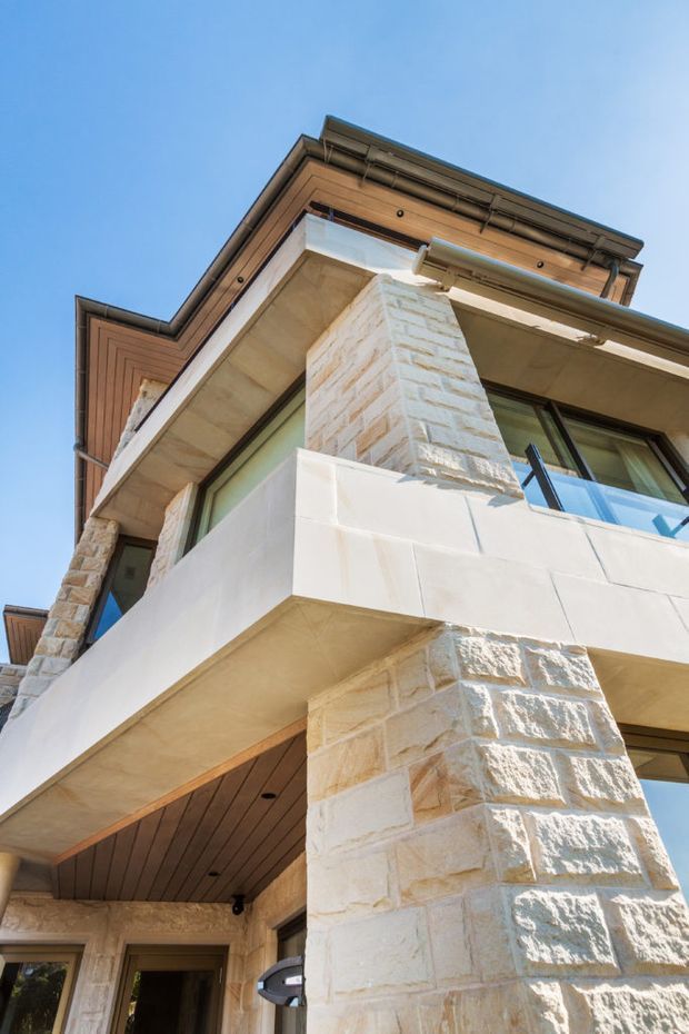 Upward-looking exterior architectural photograph of a multi-level residence with prominent pale stone masonry columns and walls, projecting balconies, dark-framed glazing, and warm timber-lined soffits and eaves. The facade is strongly articulated with layered overhangs and classical detailing against a clear blue sky.