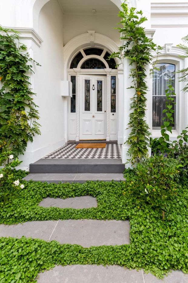 A bright, formal residential entrance viewed from the front garden. The white facade features a classical arched portico, ornate mouldings, pilasters, a white panelled front door with glazed sidelights and a fanlight, and a black-and-white checkerboard tiled porch. Dark stone steps lead through a lush garden with climbing greenery and low groundcover.