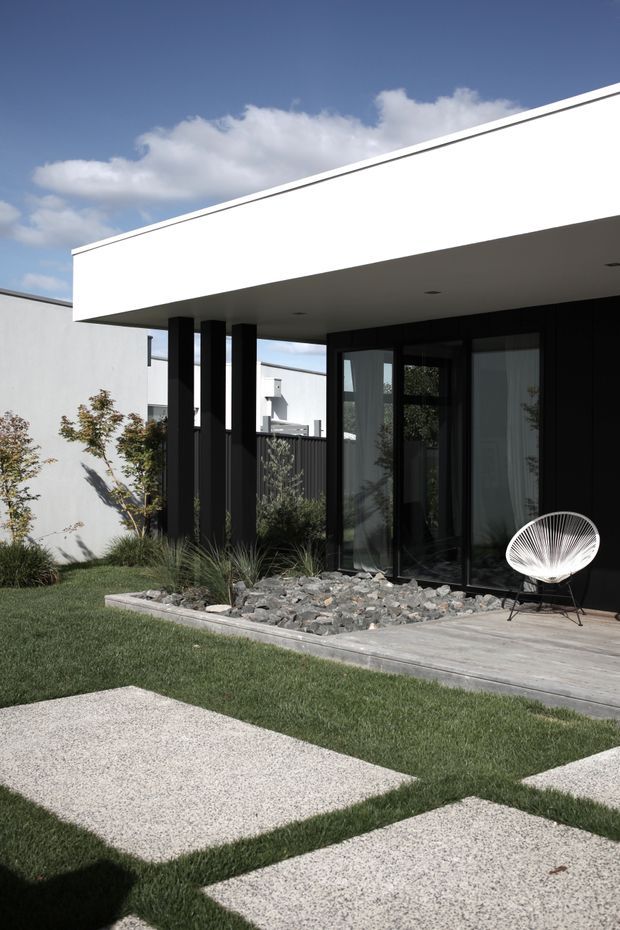 Contemporary single-storey residential exterior with a crisp white flat-roofed canopy, dark slender columns, extensive black-framed glazing, and a timber-toned deck. A minimalist landscaped garden includes gravel, grasses, small trees, lawn, and large pale concrete stepping slabs. The bright architectural composition has strong black-and-white contrast.